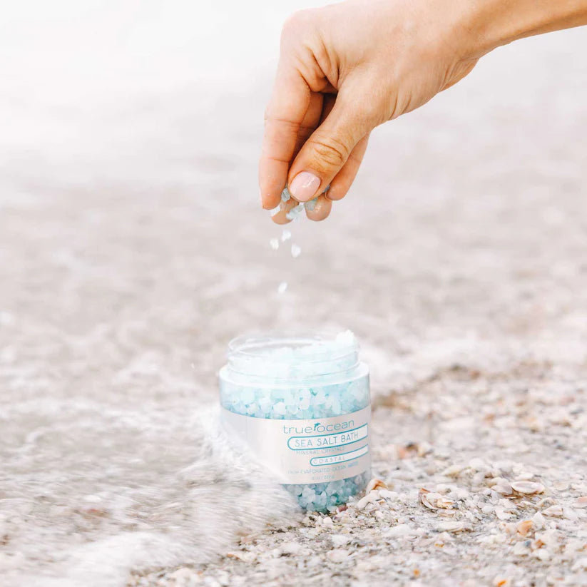 Hand sprinkling sea salt from a jar labeled 'Cruelty Free' on a sandy surface.